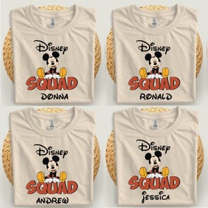 Op de afbeelding: Vier cr&egrave;mekleurige t-shirts met een Mickey Mouse-afbeelding en de tekst "Disney Squad" in het rood, met de namen Donna, Ronald, Andrew en Jessica. De shirts worden gepresenteerd op een geweven oppervlak.