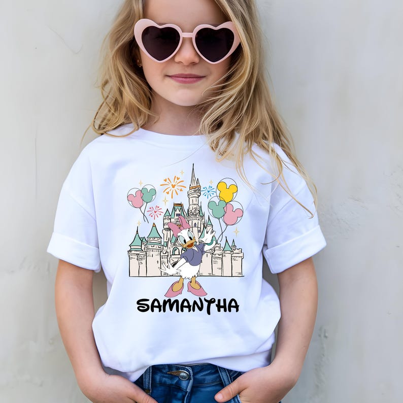 Może przedstawiać: Biała koszulka z kresk&oacute;wką Kaczki Daisy przed zamkiem, balonami i fajerwerkami. Imię "SAMANTHA" jest wydrukowane na czarno pod grafiką. Koszulka jest noszona przez osobę noszącą okulary przeciwsłoneczne w kształcie serca.