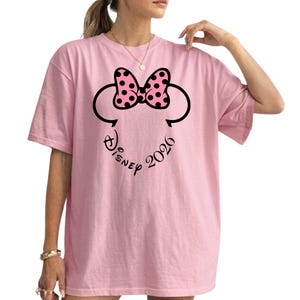 May include: A light pink t-shirt featuring a Minnie Mouse design. The design includes a black outline of Minnie's head, a pink bow with black polka dots, and the text "Disney 2026" in a script font.
