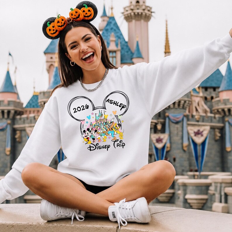 May include: White sweatshirt with a Disney Trip graphic featuring Mickey Mouse ears, a castle, and cartoon characters. The sweatshirt includes the text "2026" and "Ashley". The person is wearing a pumpkin-themed headband.