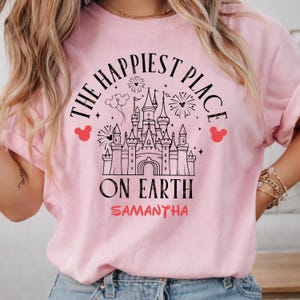 May include: A light pink sweatshirt with the text "THE HAPPIEST PLACE ON EARTH" in black, with a castle graphic and red Mickey Mouse head icons. The name "SAMANTHA" is printed in red below the text.
