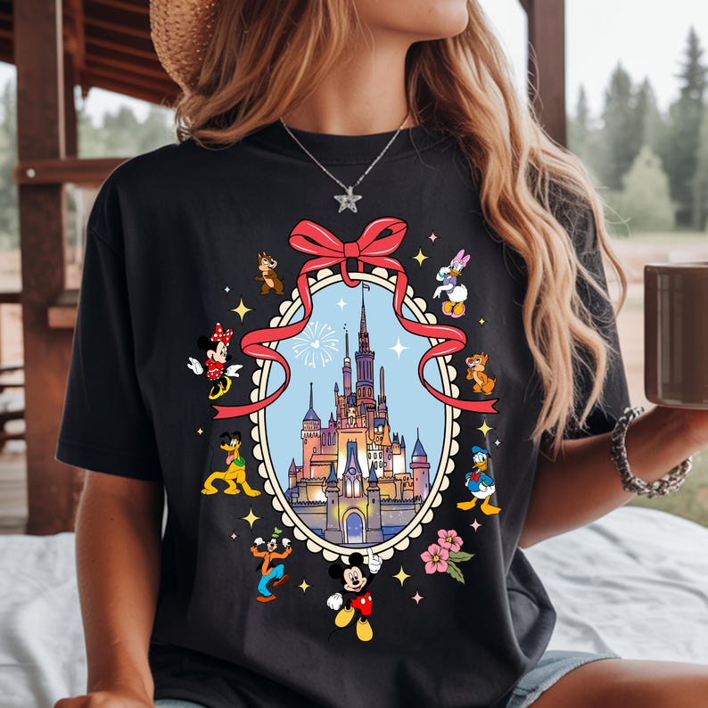 May include: Black t-shirt featuring a colorful illustration of a fairytale castle within an oval frame, surrounded by cartoon characters. A large red bow adorns the top of the frame. The design includes Mickey Mouse, Minnie Mouse, Donald Duck, and other Disney characters.