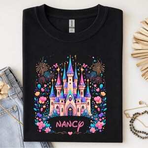 K&ouml;nnte beinhalten: Schwarzes Sweatshirt mit einem farbenfrohen Schlossdesign, Feuerwerk, Blumen und Schmetterlingen. Das Schloss hat blaue, rosa und lila T&uuml;rme. Der Name "Nancy" steht in rosa Schrift darunter.