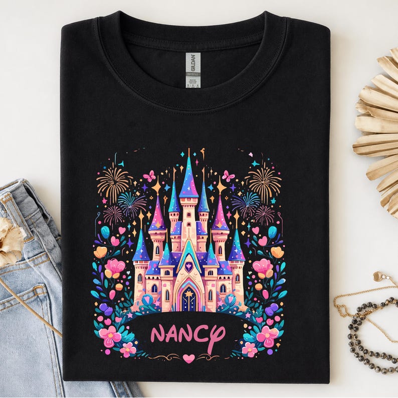 K&ouml;nnte beinhalten: Schwarzes Sweatshirt mit einem farbenfrohen Schlossdesign, Feuerwerk, Blumen und Schmetterlingen. Das Schloss hat blaue, rosa und lila T&uuml;rme. Der Name "Nancy" steht in rosa Schrift darunter.