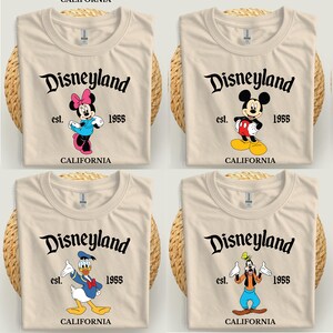 Op de afbeelding: Vier beige T-shirts met Disneyland-graphics. Elk shirt heeft een ander Disney-personage: Minnie Mouse, Mickey Mouse, Donald Duck en Goofy. De tekst "Disneyland est. 1955 CALIFORNIA" staat op elk shirt.