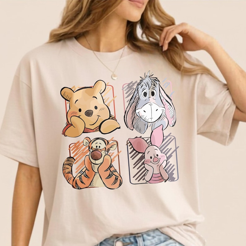 May include: Beige t-shirt featuring four cartoon character illustrations: Winnie the Pooh, Eeyore, Tigger, and Piglet. The characters are set within individual colored squares. The shirt is made of a soft, comfortable material.