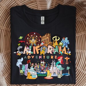May include: Black t-shirt with a vibrant graphic showcasing Disney characters and attractions. The words "CALIFORNIA ADVENTURE" are printed across the design. The image includes Mickey Mouse, a Ferris wheel, and other well-known Disney elements.
