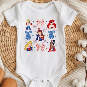 May include: White baby bodysuit with cartoon princesses in patriotic attire. The design incorporates red, white, and blue bows, fireworks, and stars. The princesses are shown in different poses, with a button fastening at the shoulder.