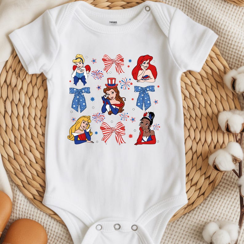 May include: White baby bodysuit with cartoon princesses in patriotic attire. The design incorporates red, white, and blue bows, fireworks, and stars. The princesses are shown in different poses, with a button fastening at the shoulder.