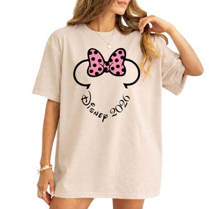May include: Beige t-shirt featuring a Minnie Mouse design. The design includes a pink polka-dot bow, black mouse ears, and the word "Disney" with the year "2026" in a curved line below the ears.