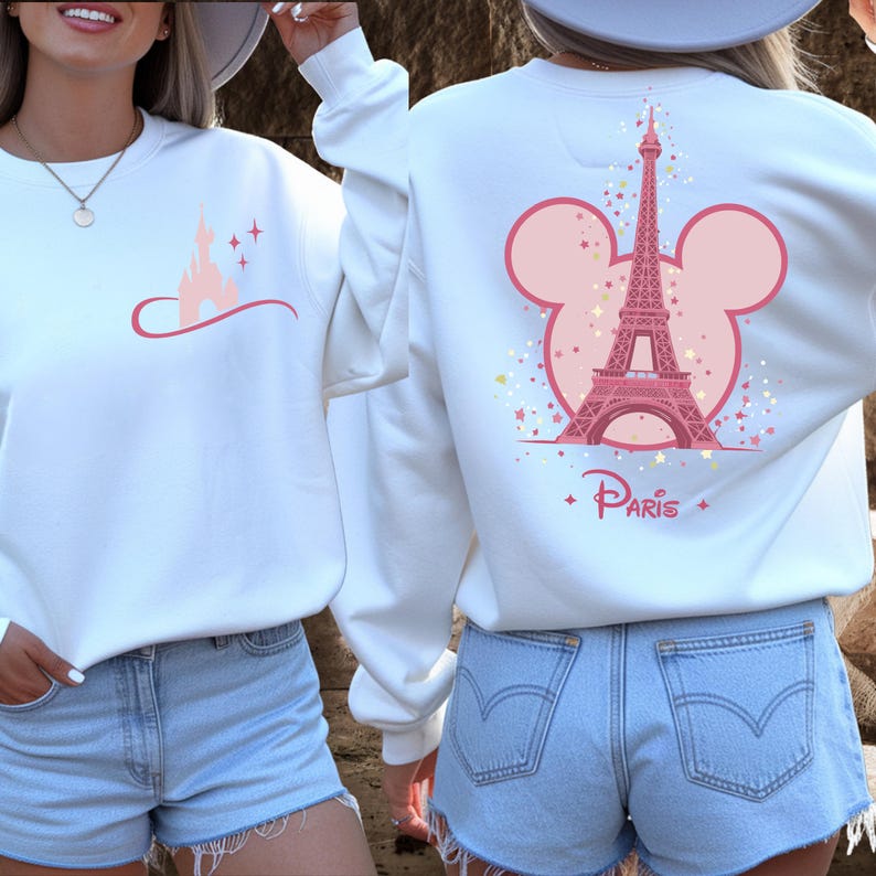 May include: White sweatshirt featuring a pink castle graphic on the front and a pink Eiffel Tower design on the back, set within a Mickey Mouse ear outline. The word "Paris" is printed below the tower.
