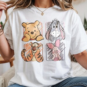 May include: White t-shirt featuring four cartoon character portraits: Winnie the Pooh, Eeyore, Tigger, and Piglet. Each character is set within a colored square, with a sketched outline. The shirt is a casual, everyday wear item.