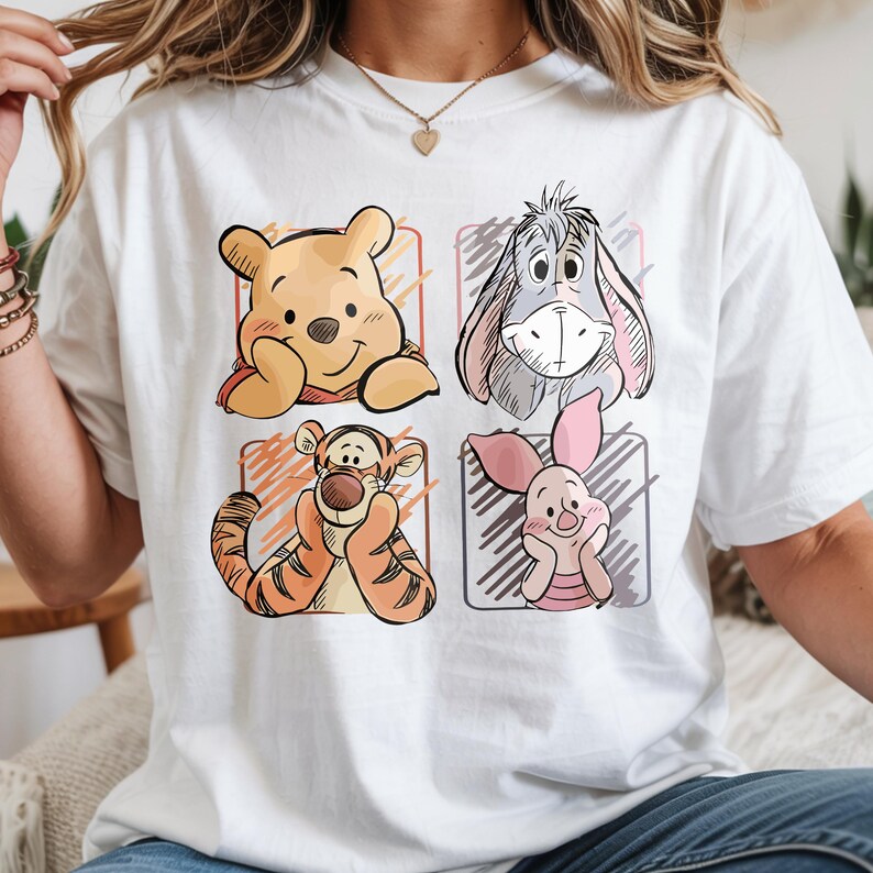 May include: White t-shirt featuring four cartoon character portraits: Winnie the Pooh, Eeyore, Tigger, and Piglet. Each character is set within a colored square, with a sketched outline. The shirt is a casual, everyday wear item.