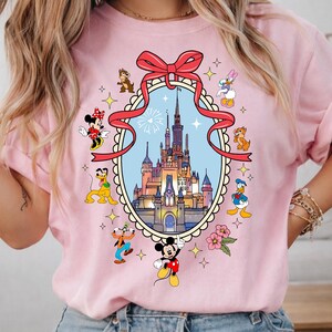May include: Pink t-shirt featuring a colorful graphic of a castle inside a decorative frame, surrounded by cartoon characters including Mickey Mouse, Minnie Mouse, Donald Duck, and Goofy. A red ribbon is at the top.