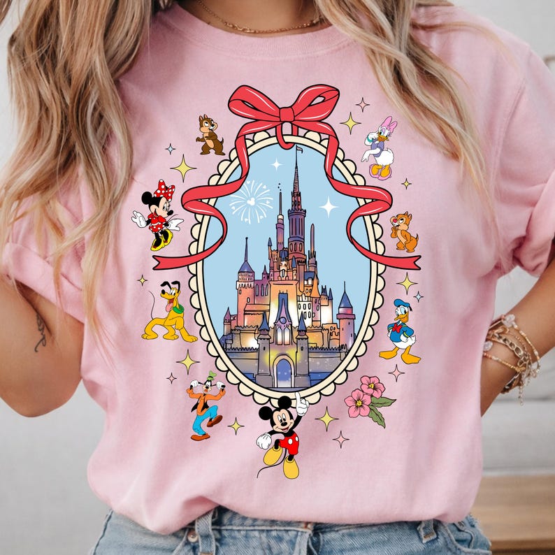 May include: Pink t-shirt featuring a colorful graphic of a castle inside a decorative frame, surrounded by cartoon characters including Mickey Mouse, Minnie Mouse, Donald Duck, and Goofy. A red ribbon is at the top.