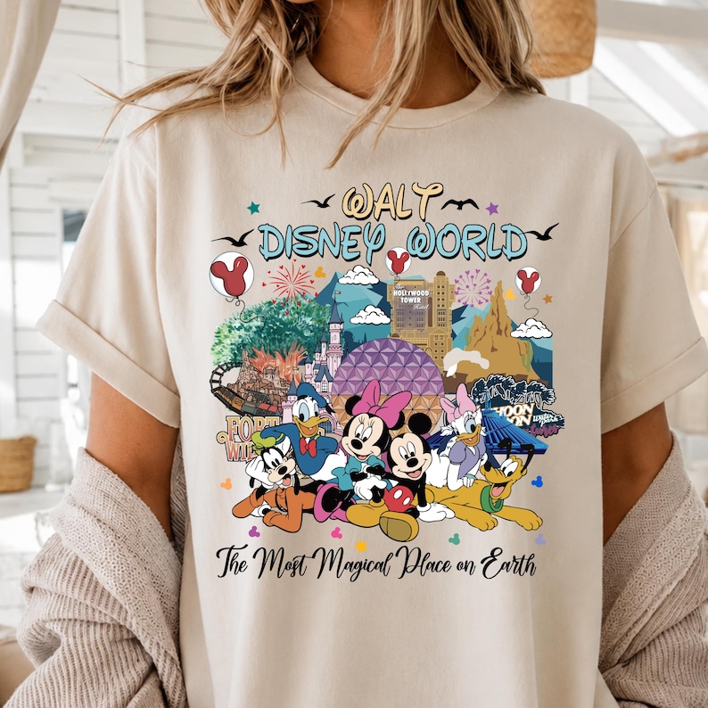 K&ouml;nnte beinhalten: Beiges T-Shirt mit einem farbenfrohen Aufdruck von Walt Disney World-Charakteren und -Sehensw&uuml;rdigkeiten. Das Design zeigt Micky und Minnie Maus, Donald Duck und Goofy, mit dem Text "Walt Disney World" und "The Most Magical Place on Earth".