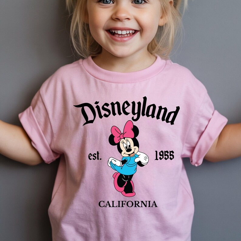 Op de afbeelding: Een lichtroze t-shirt met een Minnie Mouse-afbeelding en de tekst "Disneyland" in zwart schrift. Het shirt heeft ook de tekst "est. 1955 CALIFORNIA". De afbeelding toont Minnie Mouse in een blauwe jurk en een roze strik.