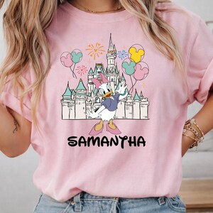 Może przedstawiać: R&oacute;żowa bluza z nadrukiem Kaczki Daisy przed zamkiem, balonami i fajerwerkami. Imię "SAMANTHA" jest wydrukowane czarnymi literami poniżej.