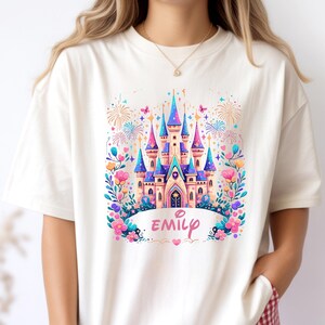 K&ouml;nnte beinhalten: Cremefarbenes T-Shirt mit einem farbenfrohen Schlossdesign in Rosa, Blau und Lila. Das Schloss ist von Blumen, Schmetterlingen und Feuerwerk umgeben. Der Name "Emily" steht in rosa Schrift darunter.