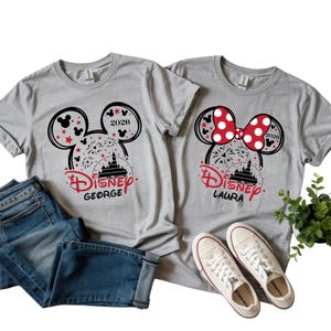 May include: Two grey t-shirts with Disney graphics. One has a Mickey Mouse head with fireworks, the text "Disney George" and "2026". The other features a Minnie Mouse bow, the text "Disney Laura" and "2026". Jeans and white trainers are also in the image.