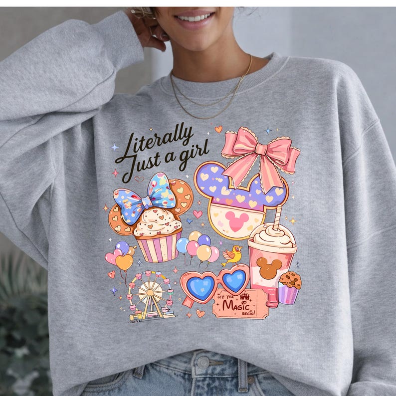 Pode incluir: Camisola cinzenta com um gr&aacute;fico colorido. Apresenta um design de orelha da Minnie Mouse, cupcakes, bal&otilde;es e uma roda gigante. O texto "Literally Just a girl" est&aacute; acima do gr&aacute;fico. Outros textos: "Magic" e "Let the... Begin!"