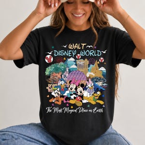K&ouml;nnte beinhalten: Schwarzes T-Shirt mit einer farbenfrohen Grafik von Disney-Figuren und Wahrzeichen. Das Design zeigt Micky und Minnie Maus, Donald Duck und Goofy, mit dem Text "Walt Disney World" und "The Most Magical Place on Earth."
