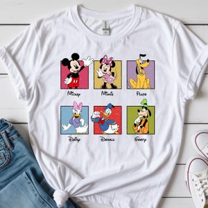 May include: White t-shirt featuring six Disney characters: Mickey, Minnie, Pluto, Daisy, Donald, and Goofy. Each character is in a colorful square with their name below. The shirt is paired with blue jeans and white sneakers.