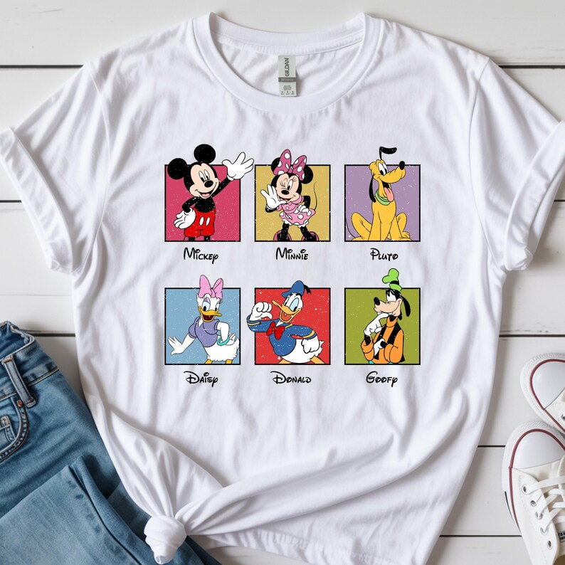 May include: White t-shirt featuring six Disney characters: Mickey, Minnie, Pluto, Daisy, Donald, and Goofy. Each character is in a colorful square with their name below. The shirt is paired with blue jeans and white sneakers.