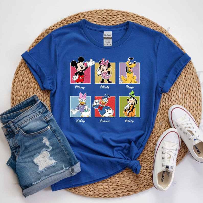 May include: Royal blue t-shirt featuring six Disney characters: Mickey, Minnie, Pluto, Daisy, Donald, and Goofy. Each character is in a colorful square with their name below. The shirt is paired with denim shorts and white sneakers.