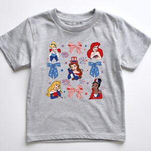 May include: A grey t-shirt with cartoon illustrations of princesses, red and white bows, and blue bows with white stars. The princesses are wearing red, white, and blue outfits. Fireworks and star graphics are also included in the design.