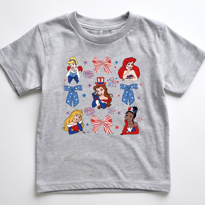 May include: A grey t-shirt with cartoon illustrations of princesses, red and white bows, and blue bows with white stars. The princesses are wearing red, white, and blue outfits. Fireworks and star graphics are also included in the design.