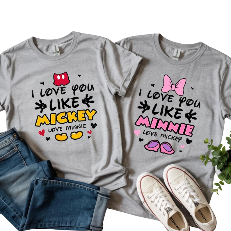 Pode incluir: Duas camisetas cinzentas com o texto "I love you like Mickey" e "I love you like Minnie" em preto, amarelo e rosa. As camisetas apresentam gr&aacute;ficos do Mickey Mouse e da Minnie Mouse, juntamente com cora&ccedil;&otilde;es e outros elementos decorativos.