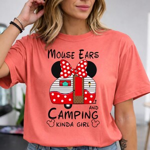 Puede incluir: Camiseta color coral con un dise&ntilde;o de caravana con forma de orejas de Minnie Mouse y un lazo rojo de lunares blancos. El texto "Mouse Ears and Camping Kinda Girl" est&aacute; escrito en negro.