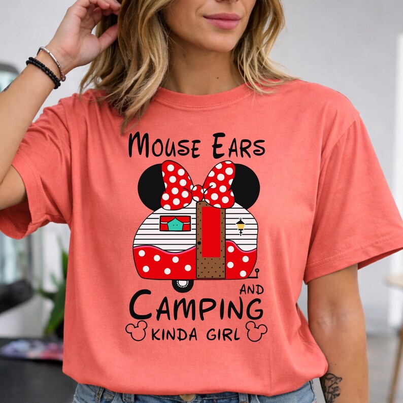 Puede incluir: Camiseta color coral con un dise&ntilde;o de caravana con forma de orejas de Minnie Mouse y un lazo rojo de lunares blancos. El texto "Mouse Ears and Camping Kinda Girl" est&aacute; escrito en negro.
