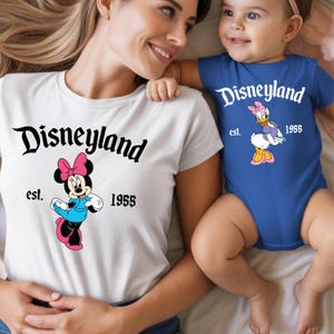Op de afbeelding: Wit T-shirt met een Minnie Mouse-afbeelding en de tekst "Disneyland est. 1955". Een blauwe babyromper met een Daisy Duck-afbeelding en de tekst "Disneyland est. 1955".