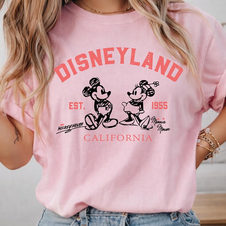 Puede incluir: Camiseta rosa claro con un dise&ntilde;o de Disneyland. El dise&ntilde;o incluye la palabra "Disneyland" en coral, junto con ilustraciones de Mickey y Minnie Mouse. Tambi&eacute;n est&aacute; presente el texto "EST. 1955" y "California".