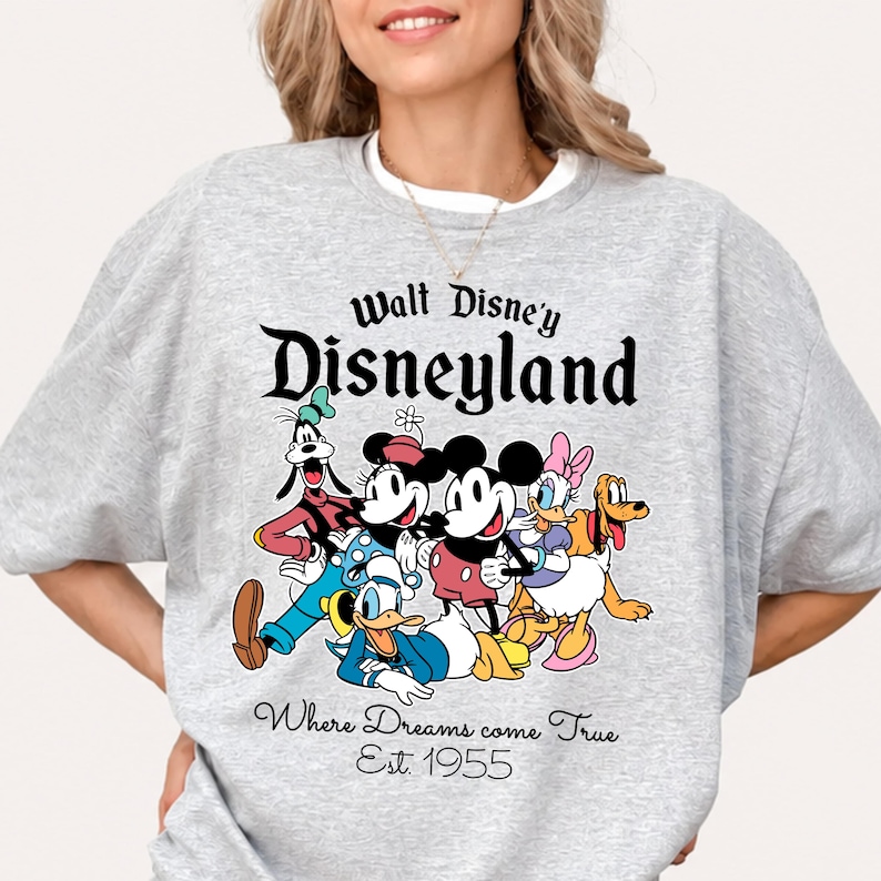 Pu&ograve; includere: Una felpa grigio chiaro con una grafica colorata dei personaggi Disney, tra cui Topolino, Minnie, Paperino e Pippo. Il testo "Walt Disney's Disneyland" e "Where Dreams come True Est. 1955" sono stampati sopra e sotto i personaggi.