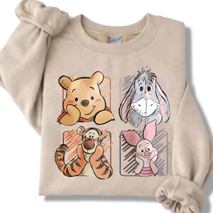 May include: Beige sweatshirt featuring four cartoon character portraits: Winnie the Pooh, Eeyore, Tigger, and Piglet. Each character is in a colored square. The sweatshirt has long sleeves and a crew neck.