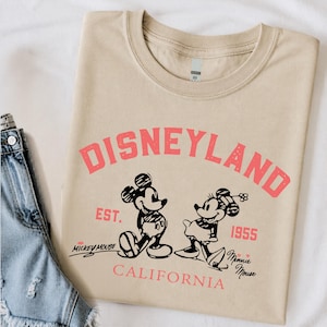 Puede incluir: Camiseta beige con la palabra "DISNEYLAND" en rosa, junto con ilustraciones de Mickey y Minnie Mouse. El texto "EST. 1955" y "CALIFORNIA" tambi&eacute;n est&aacute;n impresos en la camiseta. Una prenda cl&aacute;sica con tem&aacute;tica de Disney.