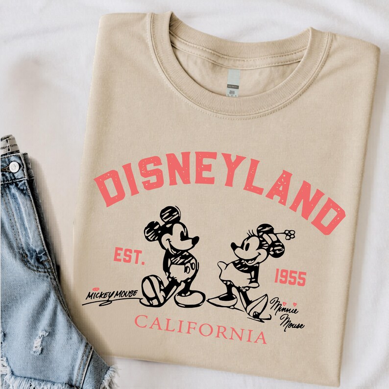 Puede incluir: Camiseta beige con la palabra "DISNEYLAND" en rosa, junto con ilustraciones de Mickey y Minnie Mouse. El texto "EST. 1955" y "CALIFORNIA" tambi&eacute;n est&aacute;n impresos en la camiseta. Una prenda cl&aacute;sica con tem&aacute;tica de Disney.