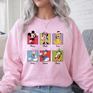 May include: A light pink sweatshirt featuring a grid of cartoon characters: Mickey, Minnie, Pluto, Daisy, Donald, and Goofy. Each character is in a colorful square with their name below. The sweatshirt is a crew neck style.