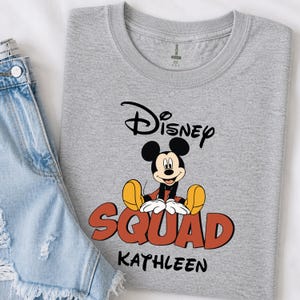 Op de afbeelding: Een grijs t-shirt met Mickey Mouse van Disney. Het shirt heeft het woord "Disney" boven Mickey en het woord "SQUAD" in oranje en zwart, met de naam "KATHLEEN" eronder. Het shirt wordt gecombineerd met een spijkerbroek.