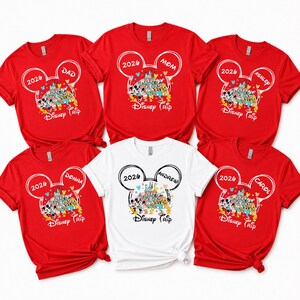 May include: Red and white t-shirts with a Disney theme. Each shirt features a Mickey Mouse head design with the year 2026 and a name, such as "Dad," "Mom," "Donna," "Andrew," "Ashley," and "Carol." The shirts have a colourful Disney castle graphic.