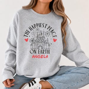 May include: A light gray sweatshirt with the text "THE HAPPIEST PLACE ON EARTH" in black, with a castle graphic and the name "ANGELA" in pink. Red Mickey Mouse head silhouettes are on either side of the text.