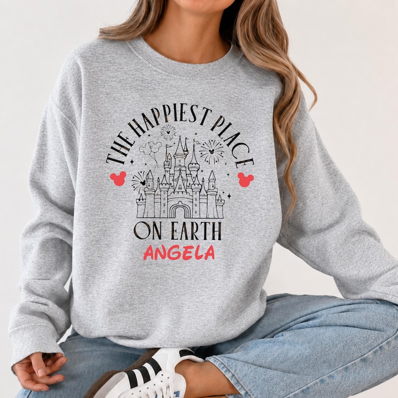 May include: A light gray sweatshirt with the text "THE HAPPIEST PLACE ON EARTH" in black, with a castle graphic and the name "ANGELA" in pink. Red Mickey Mouse head silhouettes are on either side of the text.