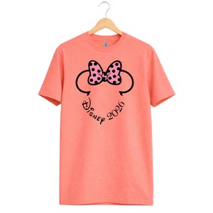 May include: Coral-colored t-shirt featuring a Minnie Mouse design. The design includes a black outline of Minnie's head, a pink bow with black polka dots, and the text "Disney 2026" in a script font. The shirt is hanging on a wooden hanger.