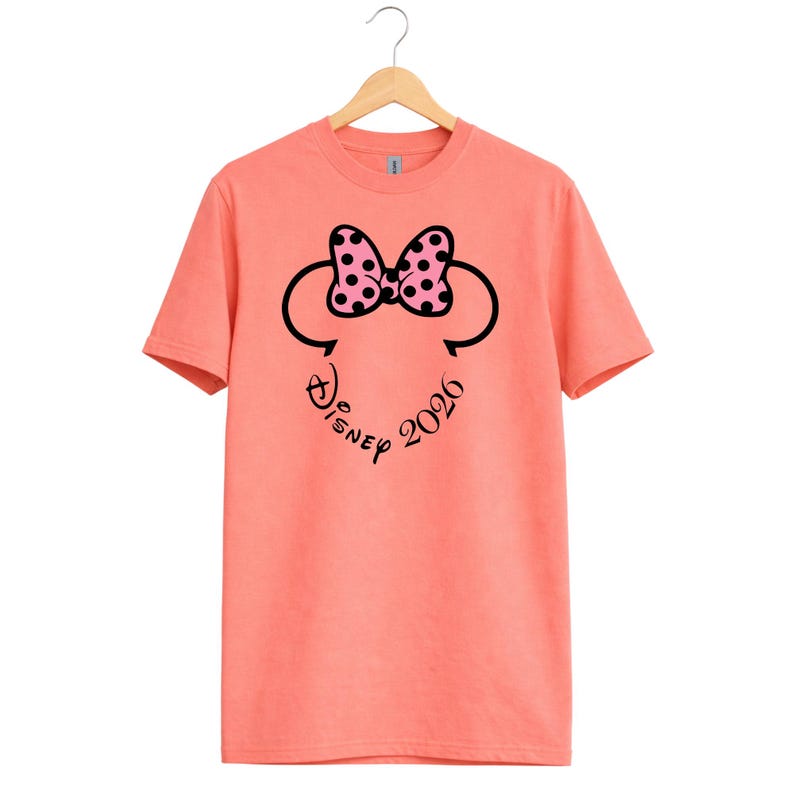 May include: Coral-colored t-shirt featuring a Minnie Mouse design. The design includes a black outline of Minnie's head, a pink bow with black polka dots, and the text "Disney 2026" in a script font. The shirt is hanging on a wooden hanger.