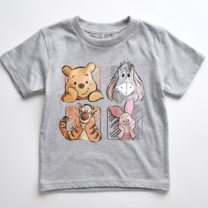 May include: A gray t-shirt featuring four cartoon character portraits: Winnie the Pooh, Eeyore, Tigger, and Piglet. The illustrations are in a sketched style, with each character in a separate square. The shirt is made of a soft, comfortable material.