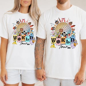 May include: Two white t-shirts with a colourful graphic design. The design includes the text "WORLD Tour 2026", landmarks, and cartoon characters. The shirts are being worn by two people.
