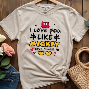 Pode incluir: Camiseta bege com o texto "I love you like Mickey loves Minnie" em preto, amarelo e vermelho. O design inclui os cal&ccedil;&otilde;es do Mickey, cora&ccedil;&otilde;es e os ic&oacute;nicos sapatos amarelos do Mickey.
